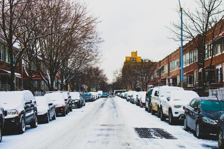SNOW COVERED ROAD NAD INLINE PARKED VEHICLES BETWEEN 2 STOREY BUILDINGS UNDER WHITE SKY